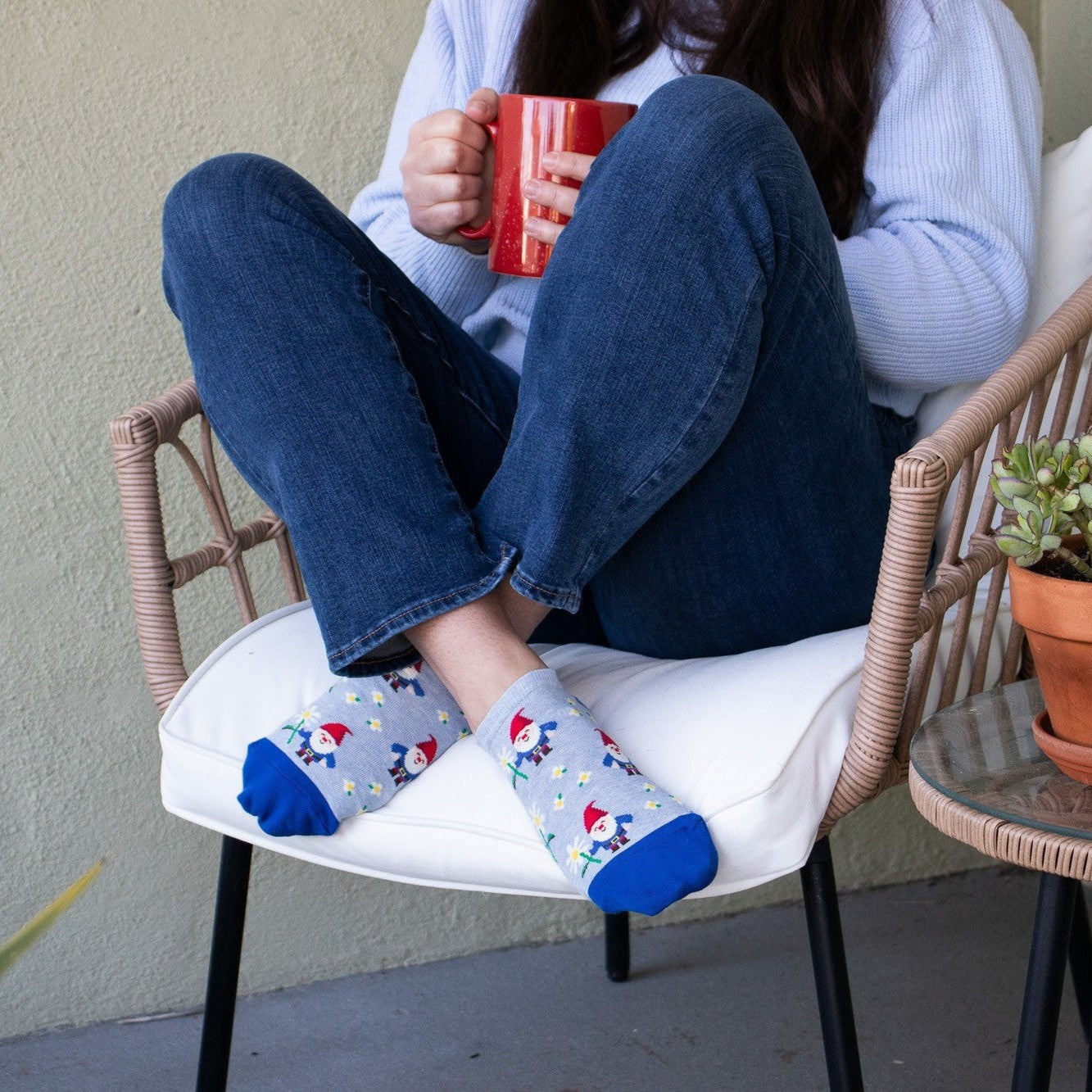A woman curls up in a wicker chair with a red mug, her crossed feet showing Growing Gnomes ankle socks in heather gray dotted with tiny gnomes and stars, paired with cuffed jeans on a sunny patio. [lf]