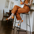 A woman in rust overalls laughs while juggling oranges from a kitchen counter stool, her Capybara Orange in Ivory socks visible on dangling feet. [lf]