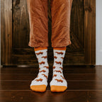 A symmetrical front view of Capybara Orange in Ivory socks worn with rust corduroy trousers on a dark hardwood floor. [lf]