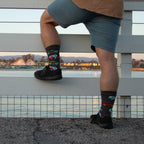 A man rests one foot on the lower rail of a white pier fence overlooking the Santa Cruz Beach Boardwalk at dusk, wearing the Woodies On The Wharf in Charcoal Heather socks printed with vintage red and teal woody wagons. [lf]