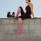 A woman in a burgundy unitard sits on the lip of a concrete skate bowl against an open blue sky, knee-high High Roller Stripe socks in Blue Heather Rain stretched up one leg as her inline skates rest beside her. [lf]