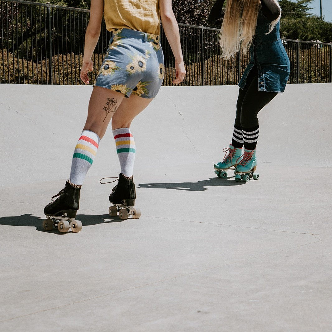 Two friends roll across a sunlit skate park, one in sunflower shorts and rainbow-striped High Roller Stripe socks in Light Gray Heather paired with black roller skates beside a partner in teal skates. [lf]