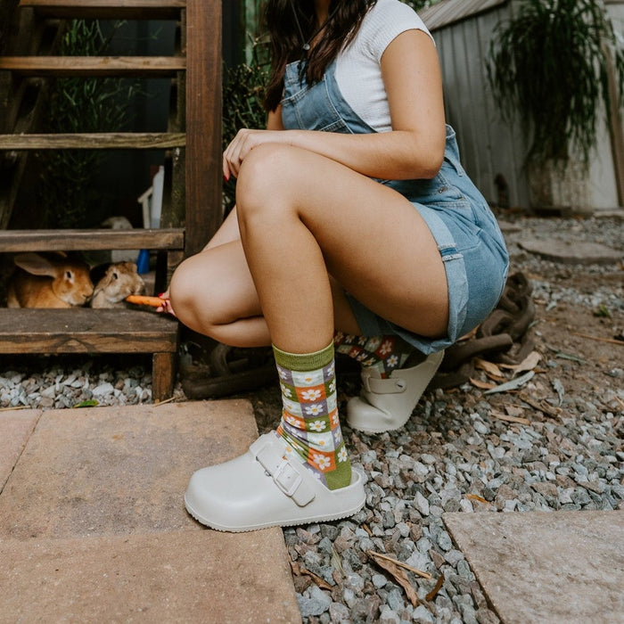 Smiling woman in a straw hat and denim overalls crouches beside a rustic wooden staircase wearing Daisy Squares Bamboo in Green socks with cream clogs. [lf]