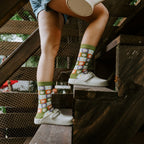 Side view of legs perched on wooden steps holding a watering can, with Daisy Squares Bamboo in Green socks tucked into cream clogs. [lf]