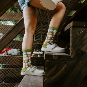 Side view of legs perched on wooden steps holding a watering can, with Daisy Squares Bamboo in Green socks tucked into cream clogs. [lf]