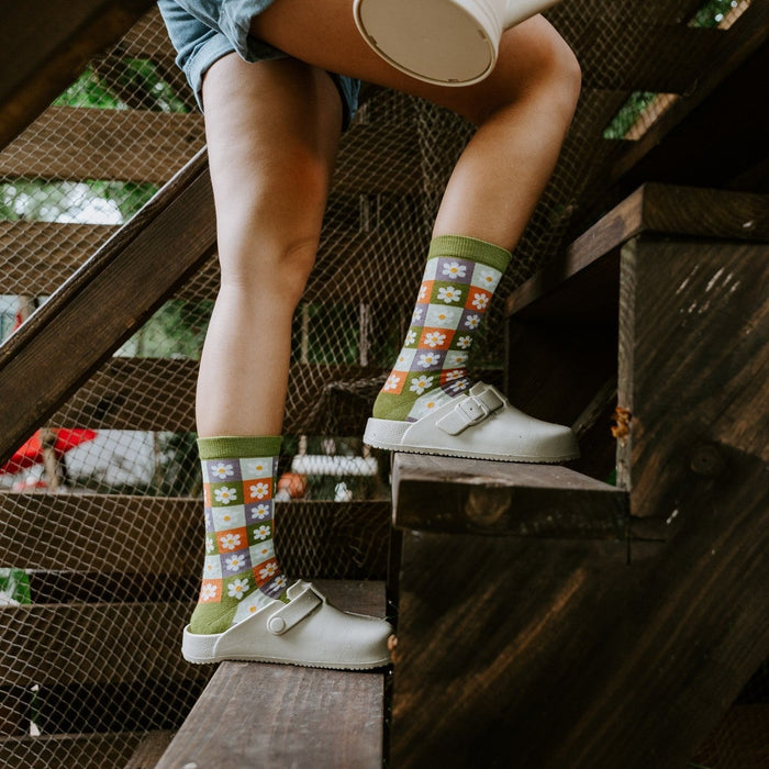 Side view of legs perched on wooden steps holding a watering can, with Daisy Squares Bamboo in Green socks tucked into cream clogs. [lf]
