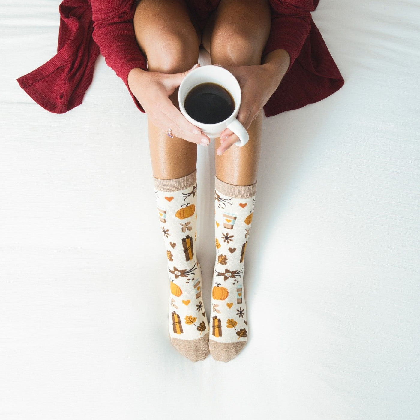 Wearer in a red sweater holds a mug of coffee with legs extended in Falling for Pumpkin Spice Bamboo socks in Ivory, pumpkin and cinnamon stick motifs across the knit. [lf]