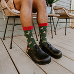 A man in a black polo and tan shorts holds a red gift box on his lap, knees together to show his green Leaping Reindeer socks and black clogs on the porch beside a potted evergreen. [lf]