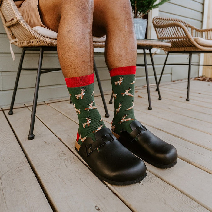 A man in a black polo and tan shorts holds a red gift box on his lap, knees together to show his green Leaping Reindeer socks and black clogs on the porch beside a potted evergreen. [lf]
