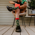 A grinning man in a reindeer-antler headband leans back in a rattan chair holding a red gift box, lifting one foot to show his green Leaping Reindeer socks and black clogs. [lf]
