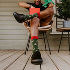 A grinning man in a reindeer-antler headband leans back in a rattan chair holding a red gift box, lifting one foot to show his green Leaping Reindeer socks and black clogs. [lf]