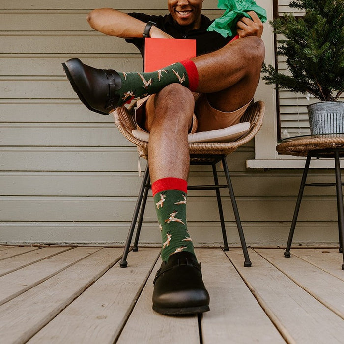 A grinning man in a reindeer-antler headband leans back in a rattan chair holding a red gift box, lifting one foot to show his green Leaping Reindeer socks and black clogs. [lf]