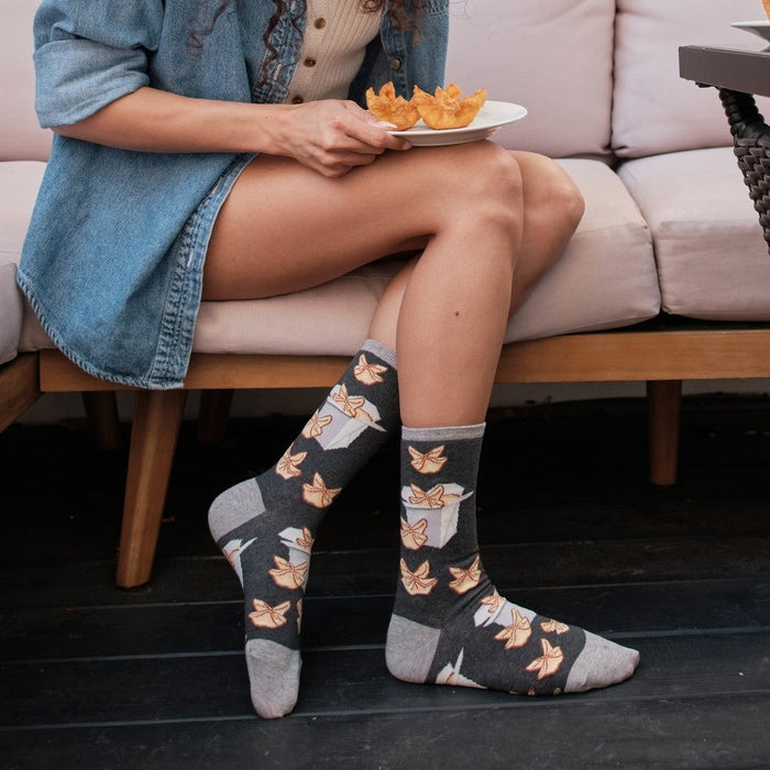 A smiling woman in a denim shirt sits on a beige patio sofa reaching toward a takeout box on a wicker side table, wearing Crab Rangoon in Charcoal Heather socks patterned with fried wontons and pagoda takeout boxes. [lf]