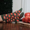 Two feet rest one over the other on a polished wood table beside a board of whole and sliced tomatoes, the green ground and red tomato print of Tantalizing Tomatoes socks in Green Heather catching window light. [lf]