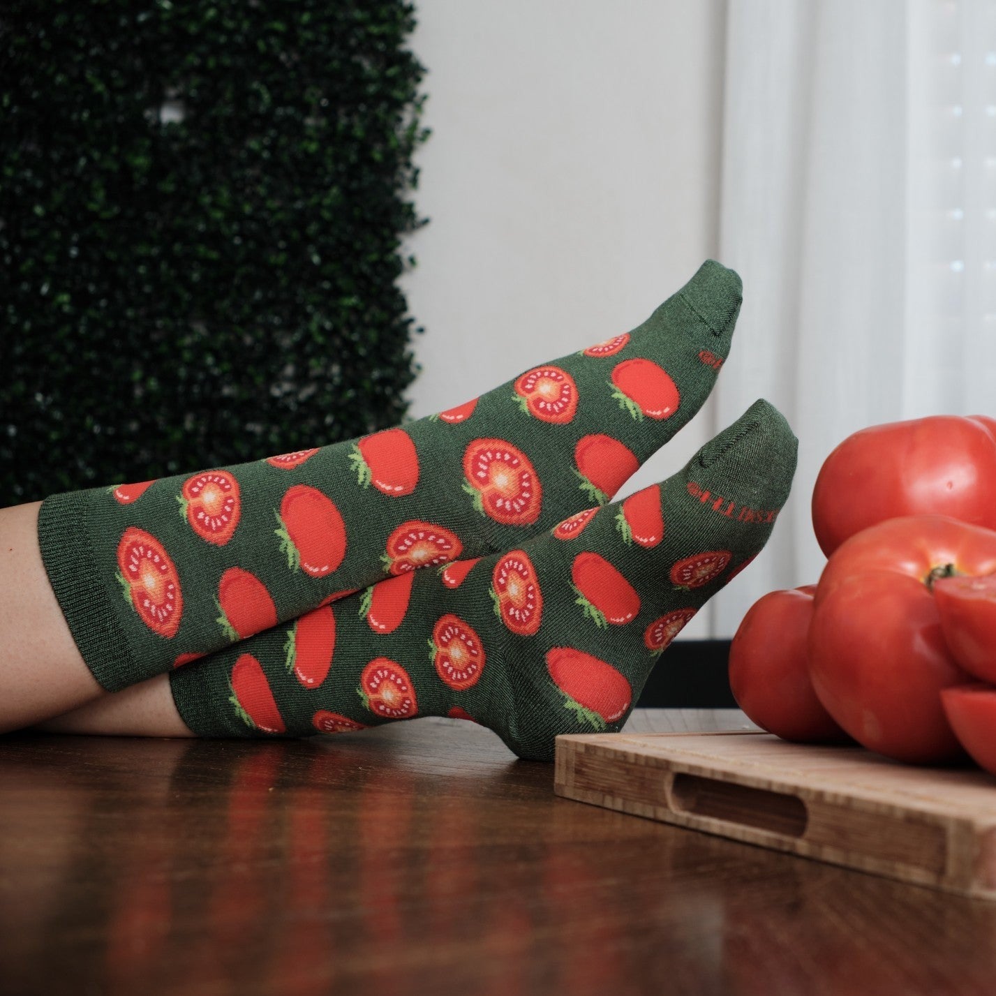 Two feet rest one over the other on a polished wood table beside a board of whole and sliced tomatoes, the green ground and red tomato print of Tantalizing Tomatoes socks in Green Heather catching window light. [lf]