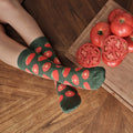 Looking down at relaxed legs on a herringbone floor next to a cutting board with tomatoes and a chef knife, the tomato print on Tantalizing Tomatoes socks in Green Heather laid out plainly from above. [lf]