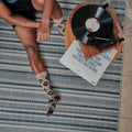 Overhead view of a man sitting on a blue and white striped rug beside a wooden record player, a vinyl LP spinning while his Vinyl Sounds Better socks in Hemp Heather show colorful record prints. [lf]