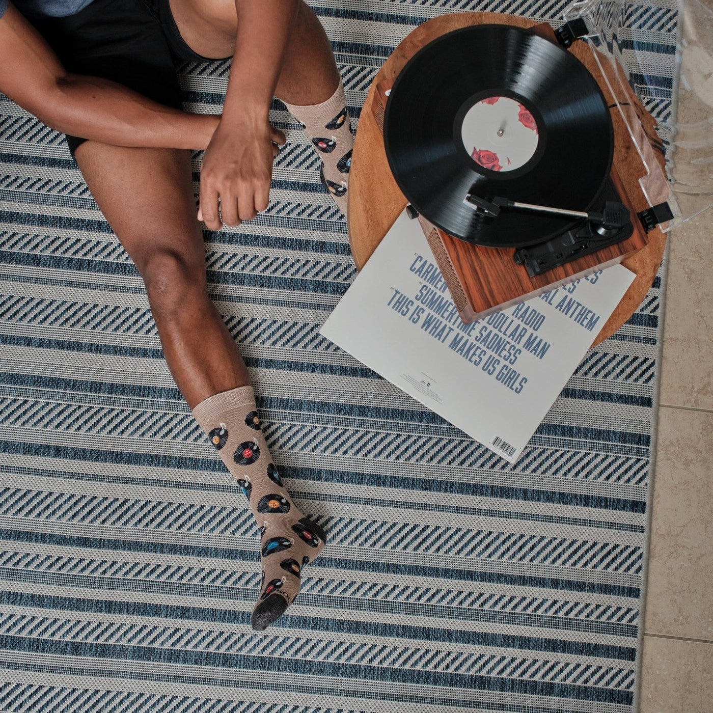 Overhead view of a man sitting on a blue and white striped rug beside a wooden record player, a vinyl LP spinning while his Vinyl Sounds Better socks in Hemp Heather show colorful record prints. [lf]