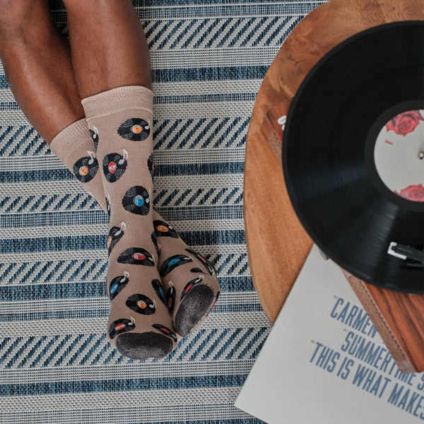 Tall overhead frame of legs propped beside a turntable and album sleeve, Vinyl Sounds Better socks in Hemp Heather catching the light of a sunlit living room. [lf]