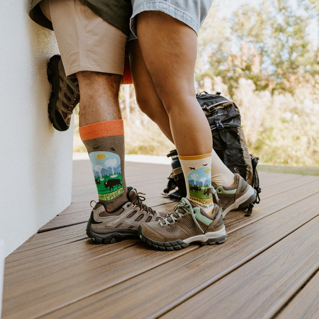 A couple in hiking boots stand close on a wood deck, his gray crew with orange cuff and her cream crew with yellow cuff both showing the Yellowstone National Park in Yellow design with bison and mountains. [lf]