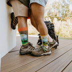 A couple in hiking boots stand close on a wood deck, his gray crew with orange cuff and her cream crew with yellow cuff both showing the Yellowstone National Park in Yellow design with bison and mountains. [lf]