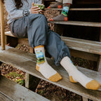 A woman sits on weathered steps cradling a green enamel mug, her cream Yellowstone National Park in Yellow socks with mountain scene resting against the wood grain. [lf]