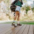 A laughing hiker mid-stride on the wooden deck adjusts her pack straps with Yellowstone National Park in Yellow socks visible between denim shorts and hiking boots. [lf]