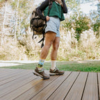 A laughing hiker mid-stride on the wooden deck adjusts her pack straps with Yellowstone National Park in Yellow socks visible between denim shorts and hiking boots. [lf]