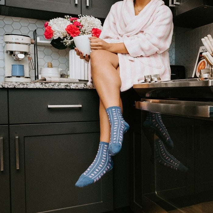 A woman in a pink robe and pink hair curlers perches on a granite kitchen counter beside a vase of flowers, swinging her feet in blue Bowquets socks above the stove. [lf]