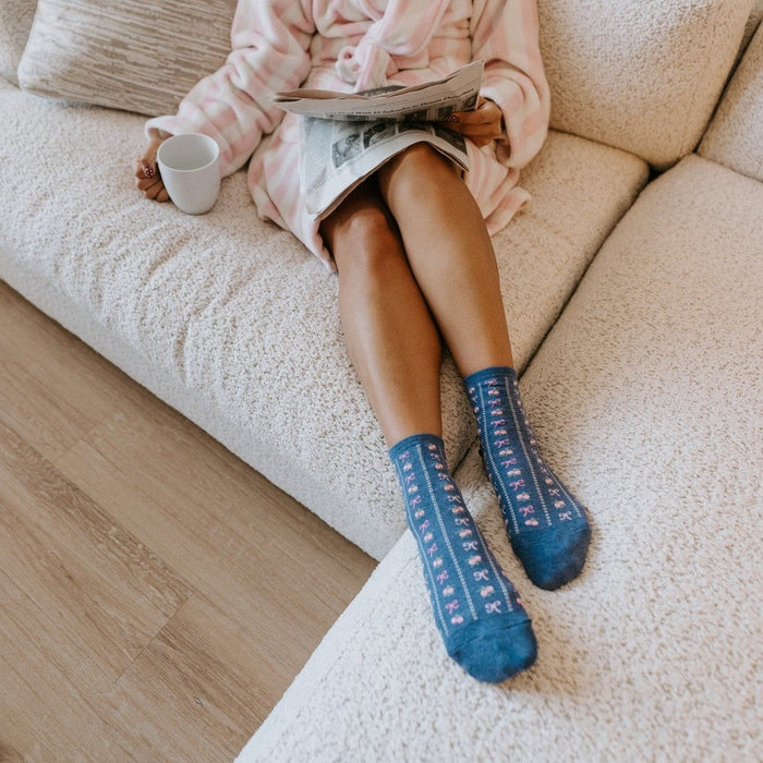 A vertical shot from above captures a woman reading a newspaper on a sectional, her blue Bowquets socks with tiny pink bows pointing toward the camera. [lf]