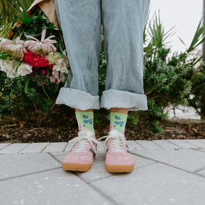 A straight-on view of feet planted on paving stones beside a hedge, showcasing Cottage Garden in Green socks with floral motifs. [lf]
