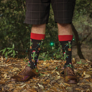 A close-up of Christmas Cactus socks in BLACK featuring Christmas cactus illustrations, on a person tending a holiday-decked plant collection. [lf]