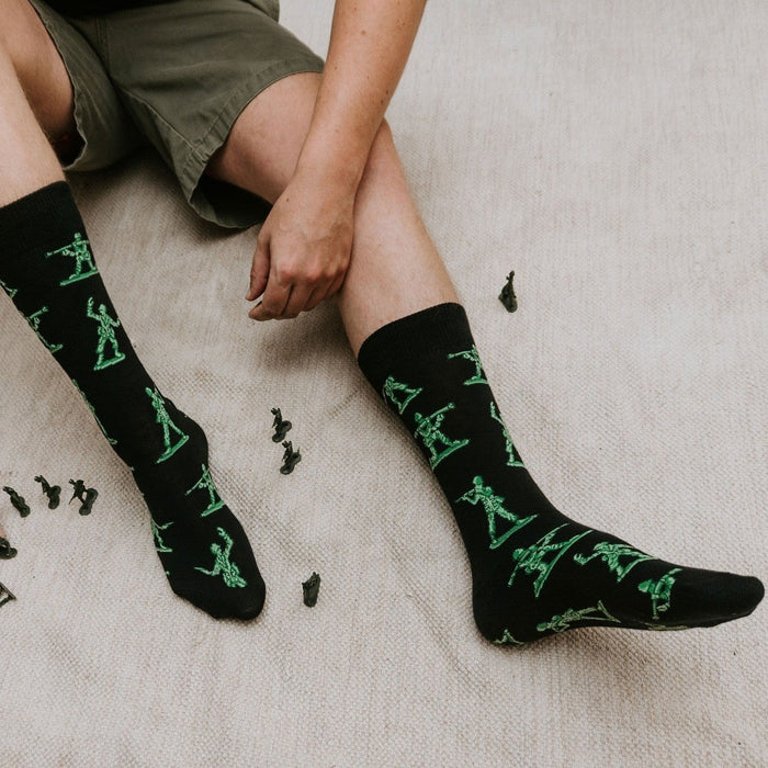 A pair of Army Men socks in BLACK stretched on a wood floor beside scattered green army figurines. [lf]