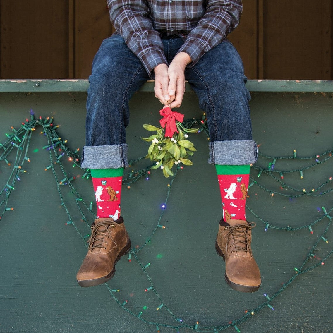 A close-up of Mythical Kissmas socks in RED featuring mistletoe and mythic creatures, on a person beside a holiday tree. [lf]