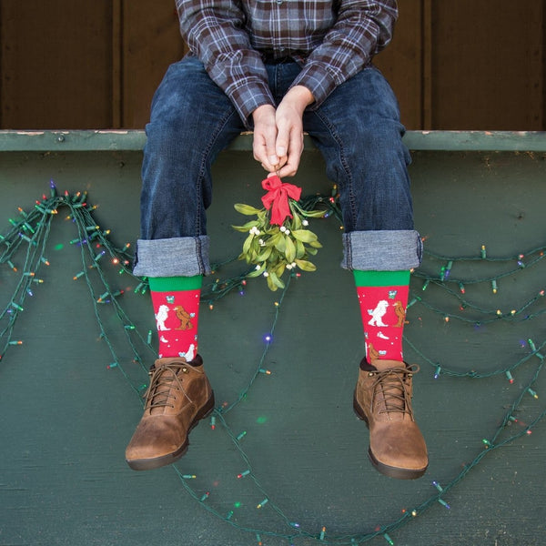 A close-up of Mythical Kissmas socks in RED featuring mistletoe and mythic creatures, on a person beside a holiday tree. [lf]