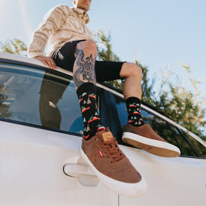 A festive overhead shot of Christmas Cars socks on a person seated on a rug beside wrapped gifts and pine garlands. [lf]