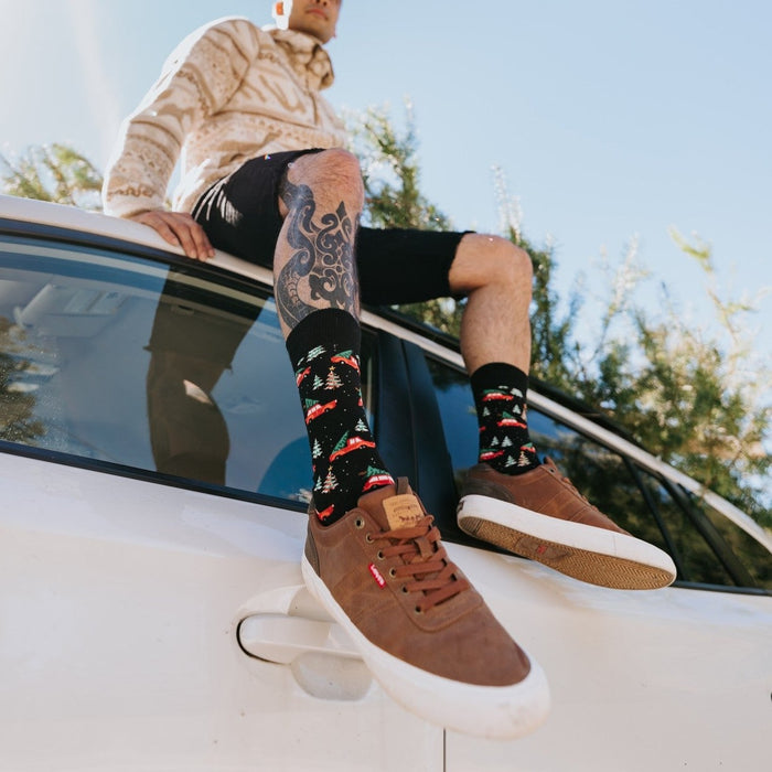 A festive overhead shot of Christmas Cars socks on a person seated on a rug beside wrapped gifts and pine garlands. [lf]