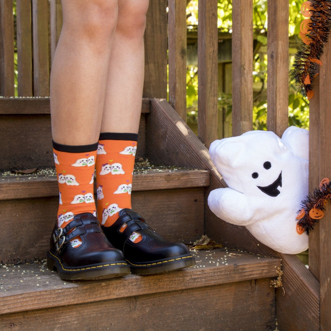 A close-up of Love You Boo socks in ORANGE featuring playful ghost illustrations with hearts, on a person on a styled Halloween sofa. [lf]
