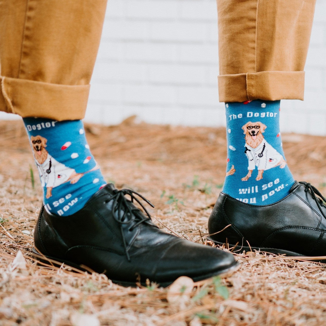 A friend in scrubs cradles a small dog, modeling The Dogtor Is In socks in Blue with playful 'dogtor' graphics. [lf]