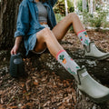 A friend in Court of Cactus socks in Light Gray Heather stands beside a tall saguaro at sunset, her socks vivid against the sand. [lf]