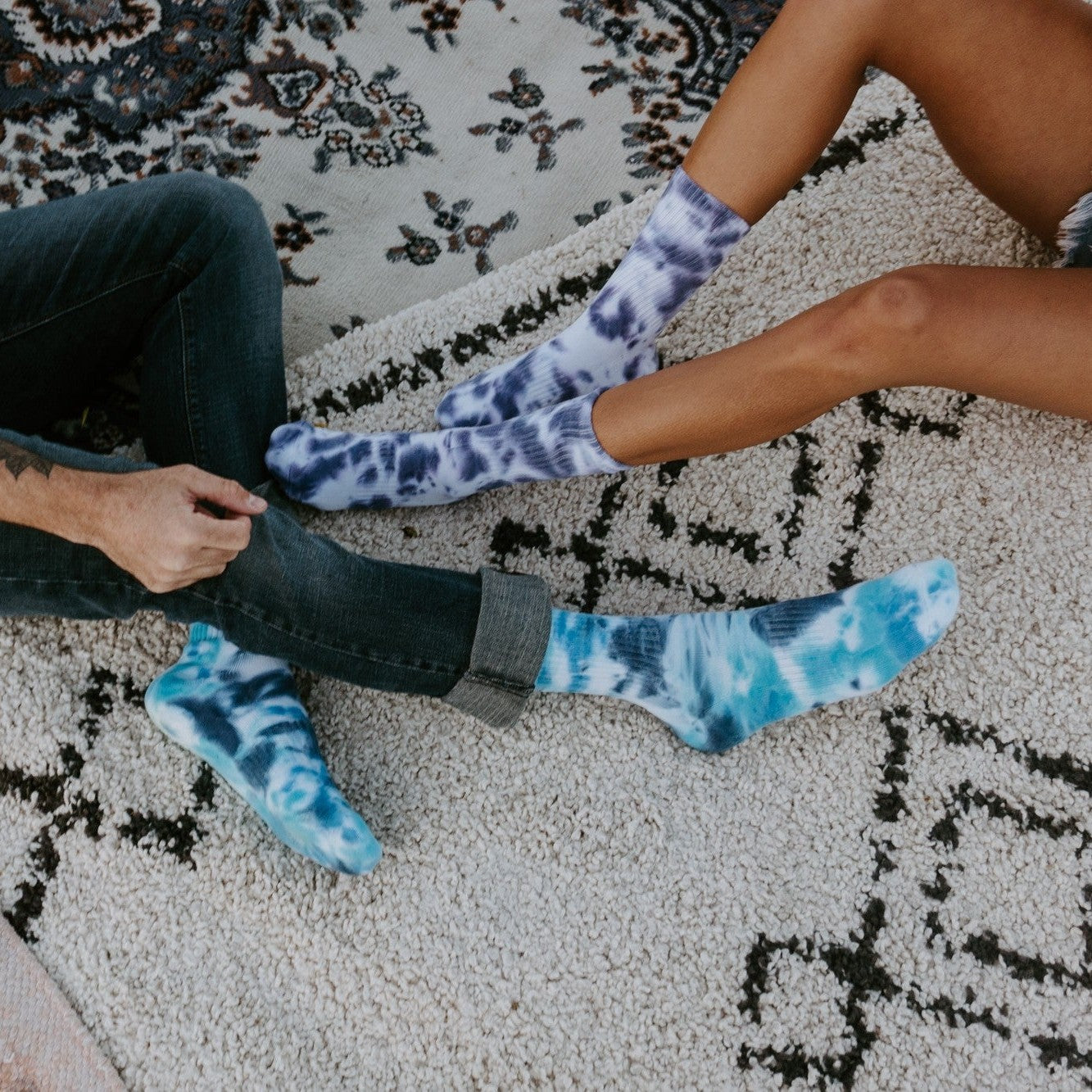 A close-up of two people seated on a patterned rug pulling on Tie Dye Athletic socks in Ink and a lighter blue variant over bare ankles. [lf]