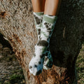 A close-up of legs draped over a tree branch with Tie Dye Athletic socks in Black Forest contrasting against the rough bark. [lf]