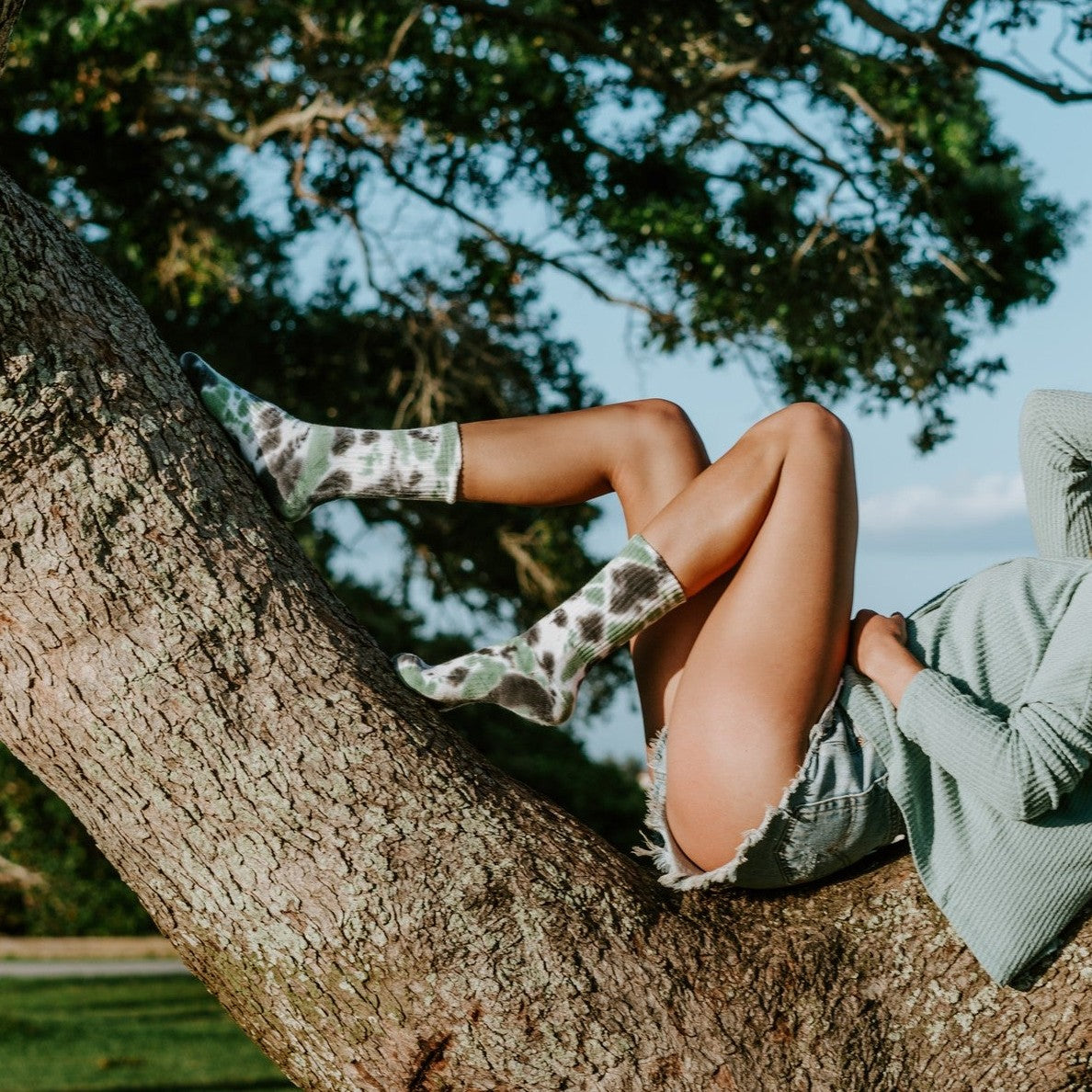 A woman in a sage long-sleeve top reclines along a sprawling tree branch with one knee up, wearing Tie Dye Athletic socks in Black Forest. [lf]