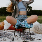 A red-haired woman in a blue crop top and denim shorts sits cross-legged on a low stool over a patterned rug, modeling Tie Dye Athletic socks in Sky Blue. [lf]