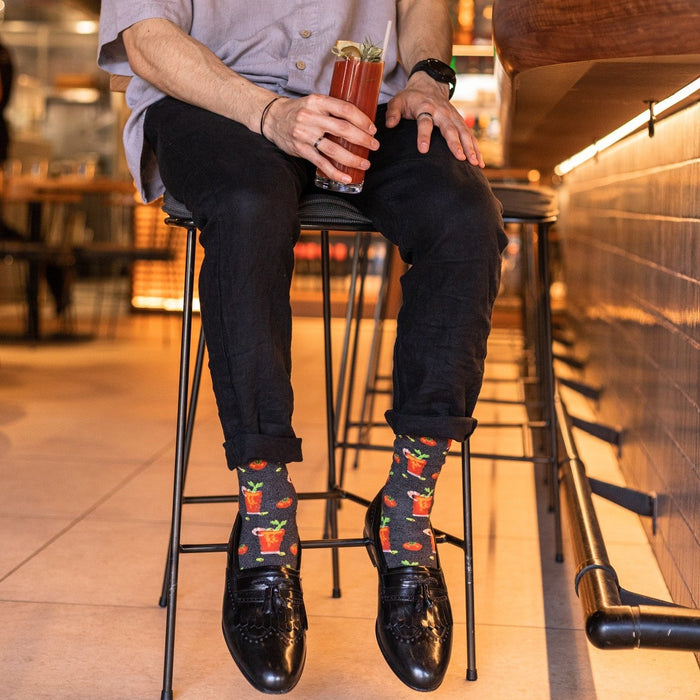 A pair of Bloody Good Drink Bamboo socks in Charcoal Heather crossed at a brunch booth beside a celery-stalk-garnished drink. [lf]