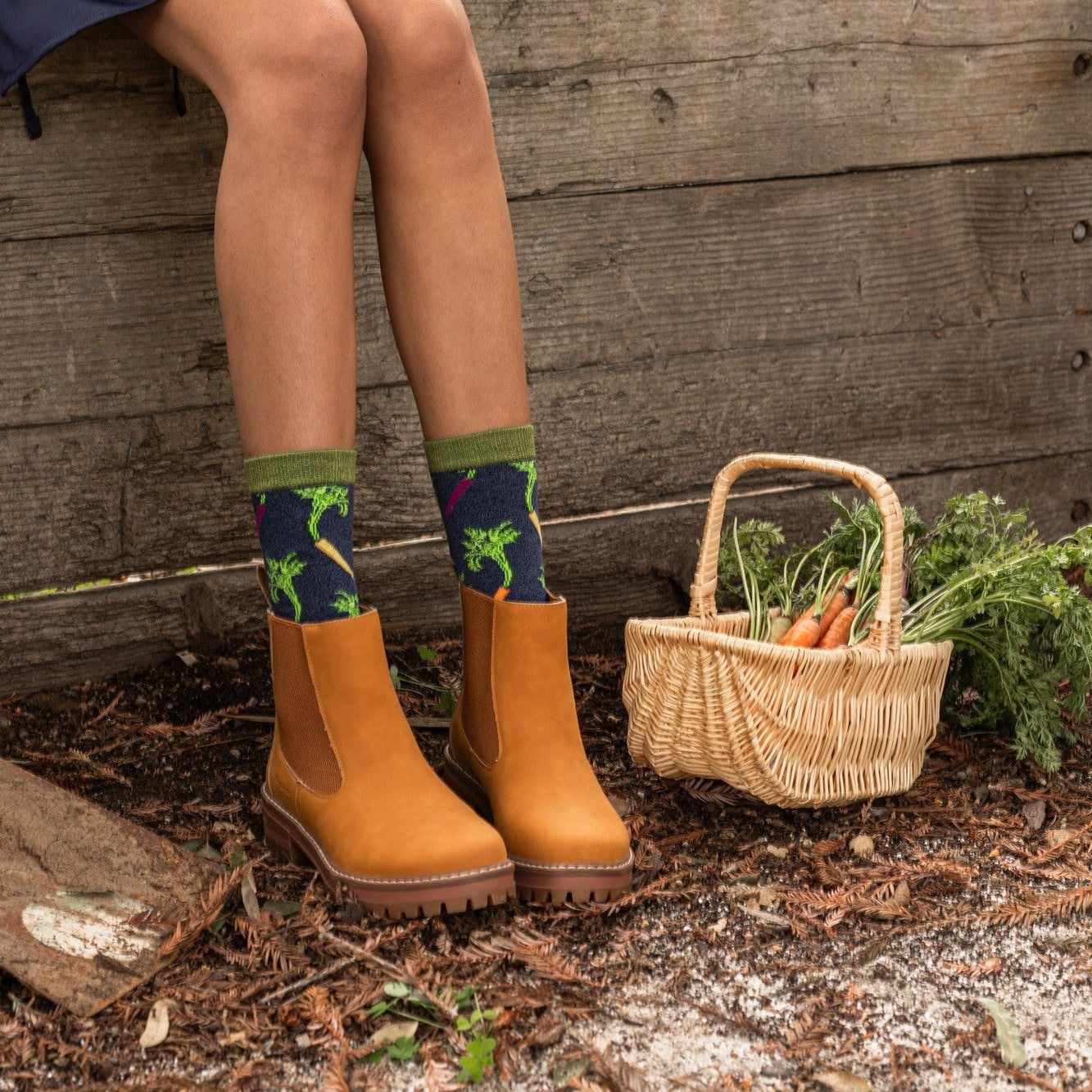 A close-up of Carrot Rainbow Bamboo socks in Navy Heather featuring rainbow carrots, on a person tending a kitchen garden. [lf]