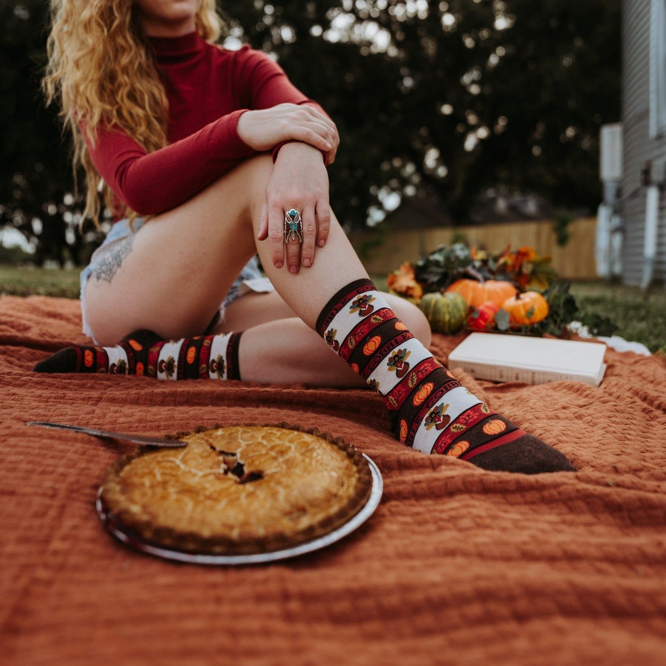 A Thanksgiving picnic flatlay with crossed feet in Gobble Gobble socks in Ivory Heather resting on a wood floor among pies, candles and autumn leaves. [lf]