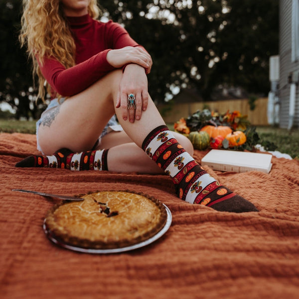 A Thanksgiving picnic flatlay with crossed feet in Gobble Gobble socks in Ivory Heather resting on a wood floor among pies, candles and autumn leaves. [lf]