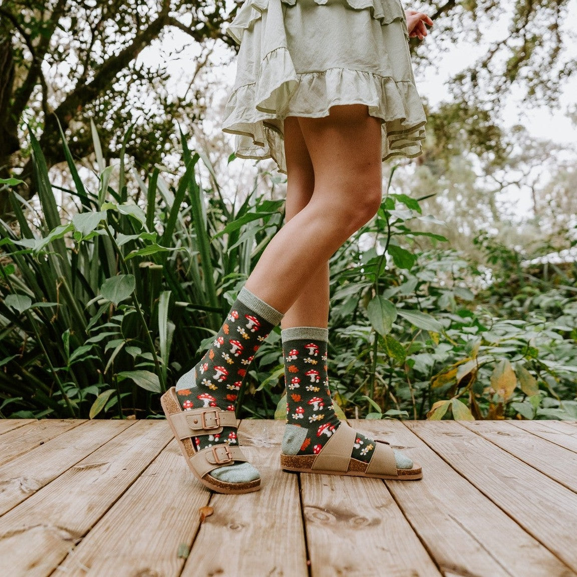 A woman in a ruffled cream dress stands on a forest deck holding wildflowers, modeling Gems Of The Forest Bamboo socks in Black with strappy brown sandals. [lf]