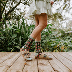 A woman in a ruffled cream dress stands on a forest deck holding wildflowers, modeling Gems Of The Forest Bamboo socks in Black with strappy brown sandals. [lf]